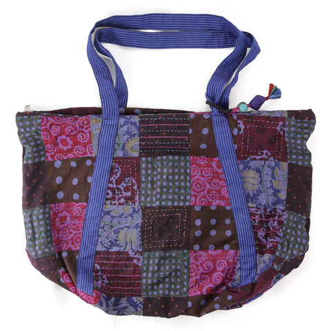 A colorful patchwork tote bag featuring a mix of patterns and colors, including red, purple, and blue squares. The bag has striped blue handles and a decorative zipper pull on top.