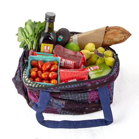A colorful cloth bag is filled with various groceries including a bottle of olive oil, fresh cherry tomatoes, radishes, an avocado, bananas, a loaf of bread, a lime, and assorted Equal Exchange packaged products.