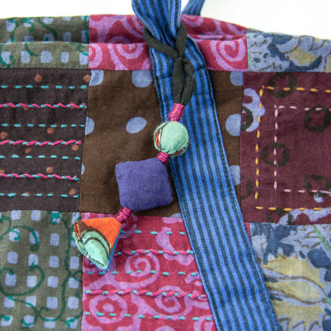 Close-up of a colorful fabric with a patchwork design featuring purple, pink, brown, and blue patches. The fabric is adorned with hand-stitched patterns and has a decorative string with fabric beads in various shapes and colors attached.
