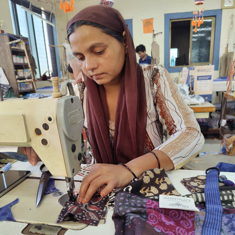A woman wearing a headscarf is focused on sewing colorful fabric pieces with a sewing machine. She is in a room with blue window frames and various crafting tools visible in the background.