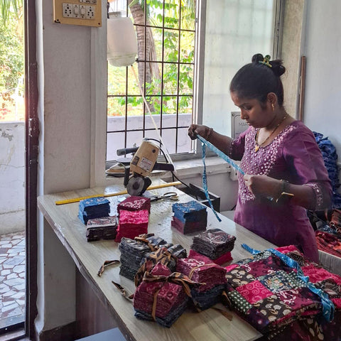 A woman wearing a purple dress is measuring ribbon at a table with stacks of colorful fabric squares. She is in a room with an open door and a window, through which greenery is visible. A sewing machine is nearby.