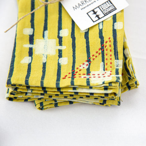 A neatly folded patterned cloth napkin with yellow and dark blue vertical stripes and white geometric designs. It has a red embroidered accent and a tag labeled Market and Equal Exchange.