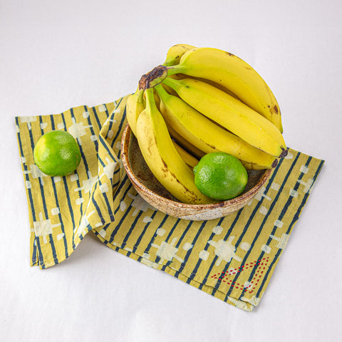 A bunch of ripe bananas in a bowl is placed on a yellow and blue patterned cloth. Two green limes accompany the bananas, one inside the bowl and one outside, on the cloth. The background is plain white.