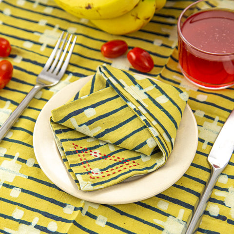 Plate with a folded yellow napkin with a geometric pattern on a matching striped tablecloth. Surrounded by a fork, knife, cherry tomatoes, bananas, and a glass of red liquid.