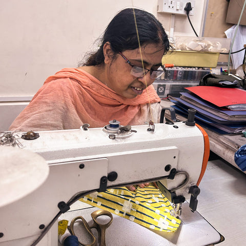 Woman with dark hair and glasses and a pink shawl using a sewing machine to sew a yellow napkin.
