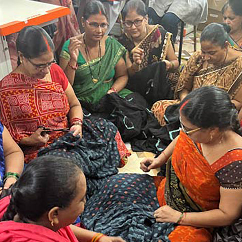 A group of women, wearing colorful sarees, sit on the floor engaged in handcrafting textiles. They appear focused, working collaboratively in a lively environment. Various fabric patterns are visible around them.