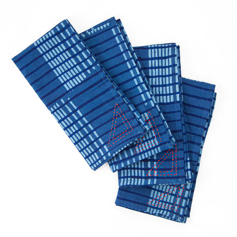 Four folded blue napkins with patterns of light blue stripes and squares, and small red stitched triangles. They are arranged in a staggered stack on a white background.