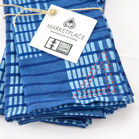 A stack of blue plaid napkins, tied with twine, featuring a stitched red triangle. A tag displays the logos for Marketplace: Handwork of India and Equal Exchange: Fairly Traded.