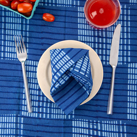 A white plate with a blue checkered napkin sits on a blue checkered napkin. A fork is on the left, and a knife is on the right. A glass of red juice is at the top, and a carton of cherry tomatoes is partially visible.