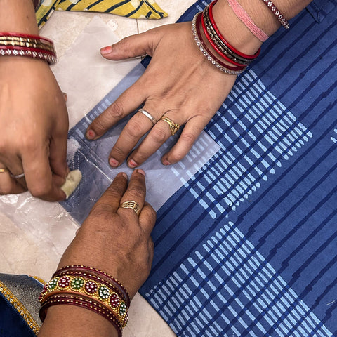 three hands wearing bracelets and rings holding a napkin, with one hand embroidering embellishment