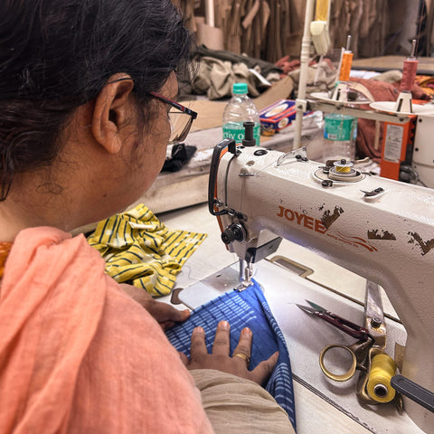 Looking over the shoulder of a woman with dark  hair and glasses using a sewing machine to sew blue napkin