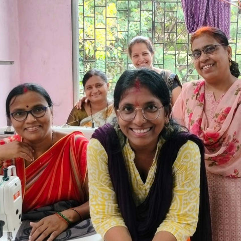 Five women smiling and posing together indoors. One is seated at a sewing machine in a red sari, and others wear casual clothing. Theyre in a room with a window showing greenery outside.
