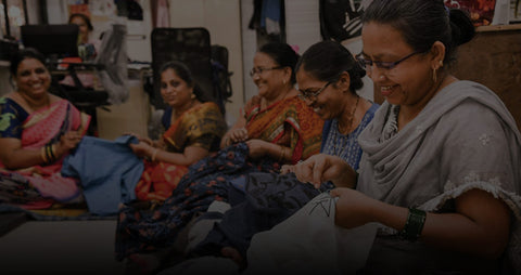 Group of five smiling women sitting on the floor embroidering colorful cloth.