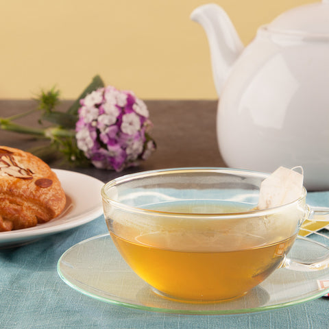 Clear cup with hot brewed chamomile tea with pastry tea pot and purple flowers in background