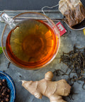 Clear cup with brewed black tea with ginger sitting on a metal tray with ginger root, a bowl of cloves, and a tea bag