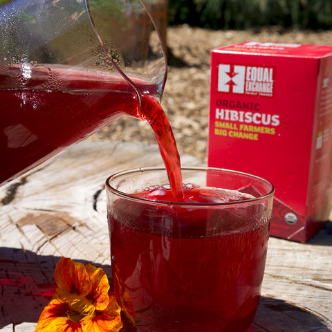 Glass pitcher pouring bright red hibiscus tea into a glass cup with a box of Equal Exchange Hibiscus tea in the sunny background