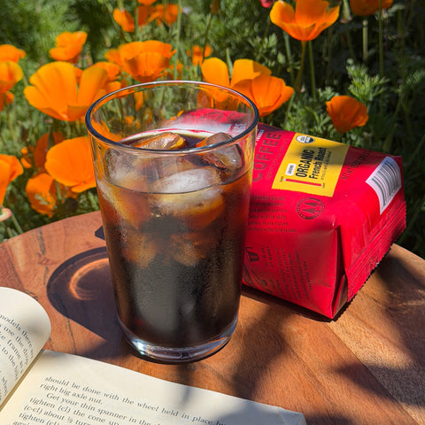 Glass of iced coffee with an Equal Exchange coffee bag and book on a wooden surface with orange flowers in the background