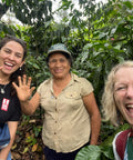 three smiling women wave to the camera in front of coffee trees