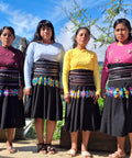 Four women coffee farmers from Chiapas standing outdoors in traditional clothing, featuring black skirts with colorful floral embroidery and wide decorative waistbands, paired with brightly colored tops.