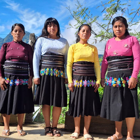 Four women coffee farmers from Chiapas standing outdoors in traditional clothing, featuring black skirts with colorful floral embroidery and wide decorative waistbands, paired with brightly colored tops.