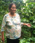 A woman standing among lush green coffee trees, wearing a light-colored floral blouse.