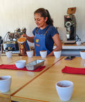 A woman in a blue apron stands behind a cupping table in a coffee quality lab, pouring hot water from a kettle into white cups. Several white ceramic cups with dark coffee are arranged on the table.