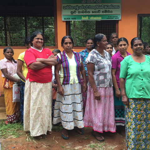 Group of SOFA tea farmers standing in a line outdoors with a building and trees in the background.