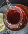 Glass teapot filled with brewed tea on a metal tray with lemon slices and loose tea leaves.