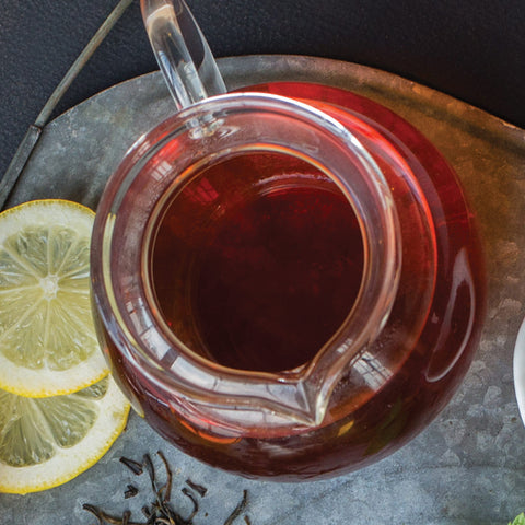 Glass teapot filled with brewed tea on a metal tray with lemon slices and loose tea leaves.