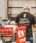 Person in a hair net and black t-shirt with "Small Farmers Big Change" packs products in warehouse with red coffee bags visible on a conveyor belt