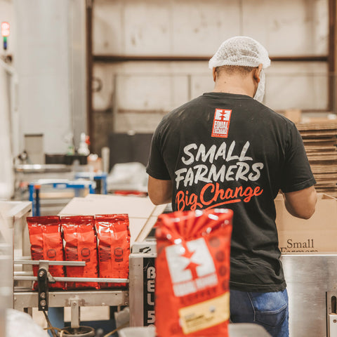 Person in a hair net and black t-shirt with "Small Farmers Big Change" packs products in warehouse with red coffee bags visible on a conveyor belt