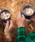 2 people holding cups of hot cocoa with mini marshmallows on a camping trip