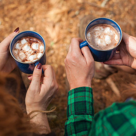 2 people holding cups of hot cocoa with mini marshmallows on a camping trip