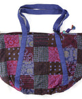 A colorful patchwork tote bag featuring a mix of patterns and colors, including red, purple, and blue squares. The bag has striped blue handles and a decorative zipper pull on top.