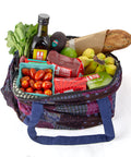 A colorful cloth bag is filled with various groceries including a bottle of olive oil, fresh cherry tomatoes, radishes, an avocado, bananas, a loaf of bread, a lime, and assorted Equal Exchange packaged products.
