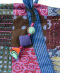 Close-up of a colorful fabric with a patchwork design featuring purple, pink, brown, and blue patches. The fabric is adorned with hand-stitched patterns and has a decorative string with fabric beads in various shapes and colors attached.