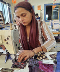 A woman wearing a headscarf is focused on sewing colorful fabric pieces with a sewing machine. She is in a room with blue window frames and various crafting tools visible in the background.