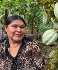 A woman wearing a leopard-print blouse stands in a lush green cocoa farm next to a tree trunk bearing large green cocoa pods.