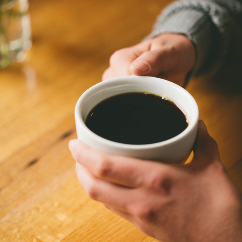 Hands holding a mug of dark black coffee at a wood table