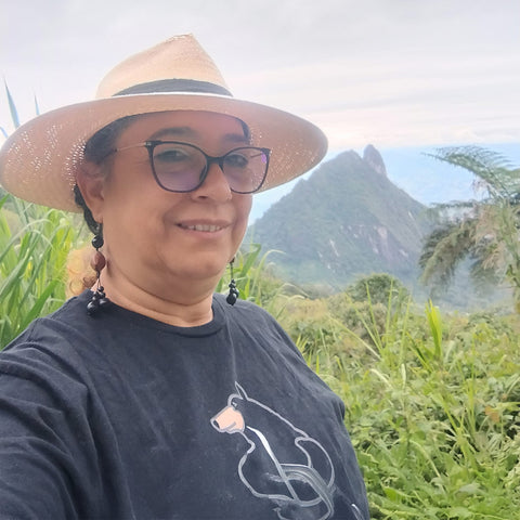 Luz Marina wearing a wide-brimmed hat and glasses, standing in a lush green landscape with mountains in the background.
