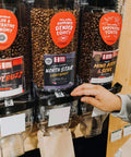 Bulk coffee bins with social impact stickers, with a hand reaching out to fill a bulk bag.