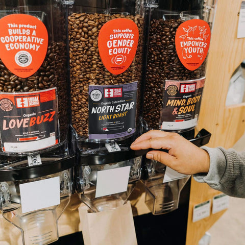 Bulk coffee bins with social impact stickers, with a hand reaching out to fill a bulk bag.