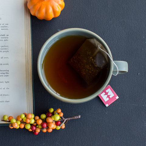 Cup of Organic Chai with tea bag steeping sitting on grey background with a book, pepper corns, and a pumpkin