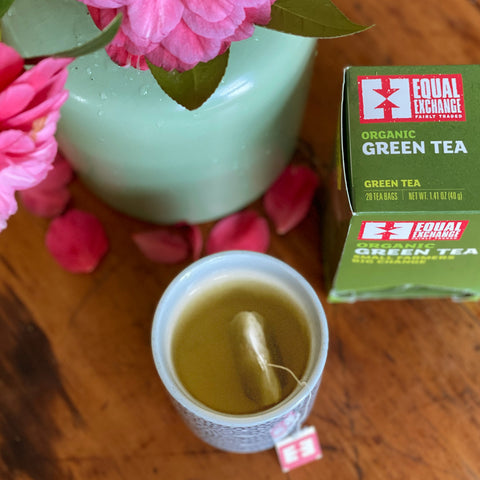 Cup of steeping green tea next to box of Equal Exchange Green Tea and a light green vase with pink flowers 