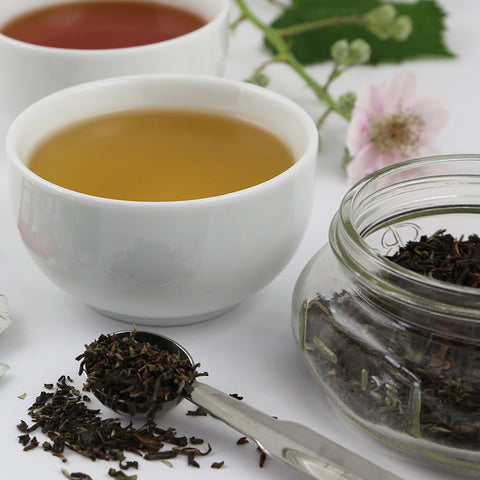 Cup of green tea next to a glass jar and scoop with loose tea leaves in front and a pink flower in the background