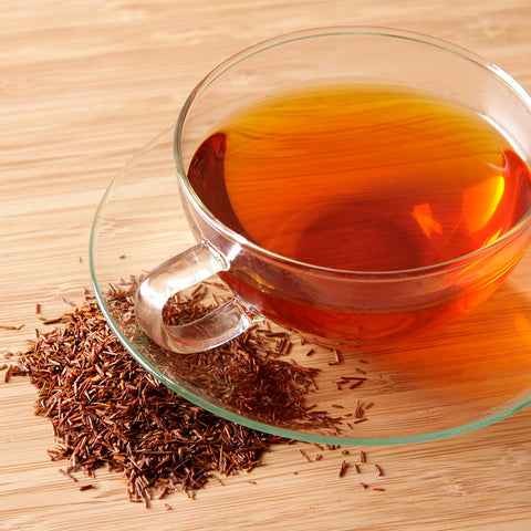 Clear cup of brewed rooibos tea with loose tea scattered in front on a wooden tabletop