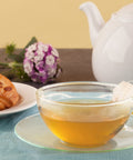Clear cup with hot brewed chamomile tea with pastry tea pot and purple flowers in background