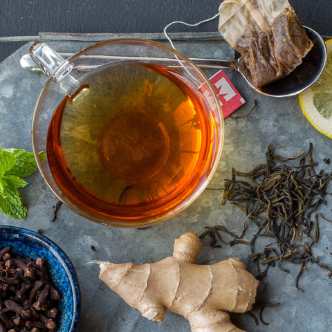 Clear cup with brewed black tea with ginger sitting on a metal tray with ginger root, a bowl of cloves, and a tea bag