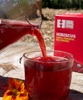 Glass pitcher pouring bright red hibiscus tea into a glass cup with a box of Equal Exchange Hibiscus tea in the sunny background