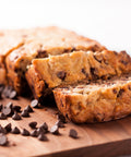 Loaf of chocolate chip banana bread sliced on a cutting board with chocolate chips scattered in front